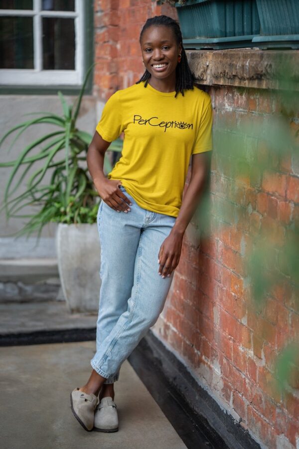 A woman leaning against a brick wall wearing jeans and a yellow shirt.